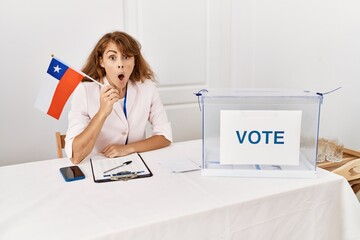 Wall Mural - Beautiful caucasian woman at political campaign election holding chile flag scared and amazed with open mouth for surprise, disbelief face