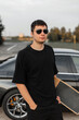 © alones - Cool man model hipster in fashion black t-shirt with longboard stands near a car outdoors