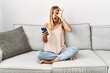 © Krakenimages.com - Blonde beautiful young woman sitting on the sofa at home using smartphone doing ok gesture shocked with surprised face, eye looking through fingers. unbelieving expression.