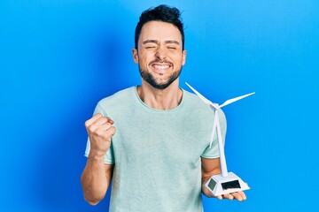 Young hispanic man holding solar windmill for renewable electricity screaming proud, celebrating victory and success very excited with raised arm