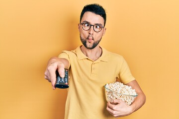 Poster - Young hispanic man eating popcorn using tv control making fish face with mouth and squinting eyes, crazy and comical.