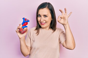 Poster - Middle age hispanic woman holding heart organ doing ok sign with fingers, smiling friendly gesturing excellent symbol