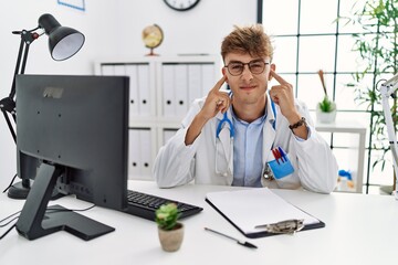 Sticker - Young caucasian doctor man working at the clinic covering ears with fingers with annoyed expression for the noise of loud music. deaf concept.