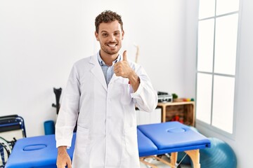 Wall Mural - Handsome young man working at pain recovery clinic doing happy thumbs up gesture with hand. approving expression looking at the camera showing success.