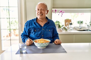 Canvas Print - Senior man with grey hair eating pasta spaghetti at home winking looking at the camera with sexy expression, cheerful and happy face.