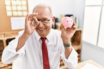 Canvas Print - Senior business man holding piggy bank smiling happy doing ok sign with hand on eye looking through fingers