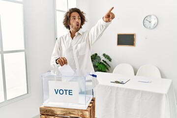 Sticker - Young hispanic man voting putting envelop in ballot box pointing with finger surprised ahead, open mouth amazed expression, something on the front