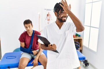 Wall Mural - Young hispanic man working at pain recovery clinic with a man with broken arm surprised with hand on head for mistake, remember error. forgot, bad memory concept.