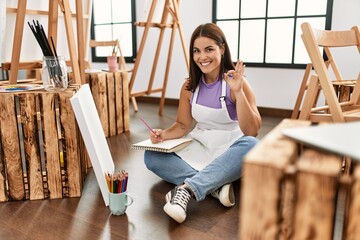 Sticker - Young beautiful hispanic woman at art studio doing ok sign with fingers, smiling friendly gesturing excellent symbol