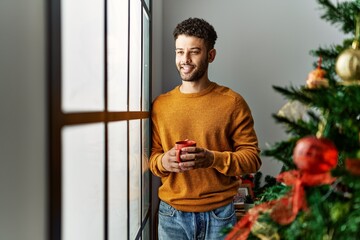 Canvas Print - Young arab man drinking coffee standing by christmas tree at home.