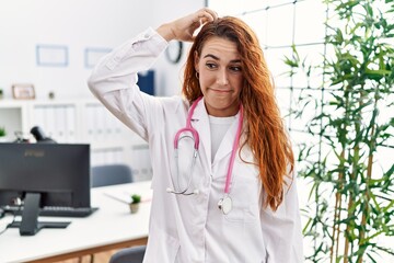 Poster - Young redhead woman wearing doctor uniform and stethoscope at the clinic confuse and wonder about question. uncertain with doubt, thinking with hand on head. pensive concept.