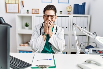 Sticker - Young man with beard wearing doctor uniform and stethoscope at the clinic laughing and embarrassed giggle covering mouth with hands, gossip and scandal concept