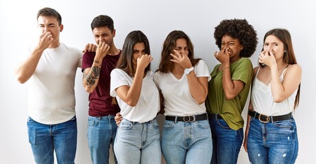 Poster - Group of young friends standing together over isolated background smelling something stinky and disgusting, intolerable smell, holding breath with fingers on nose. bad smell