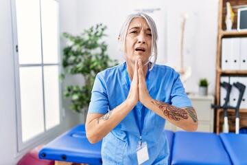 Wall Mural - Middle age grey-haired woman wearing physiotherapist uniform at medical clinic begging and praying with hands together with hope expression on face very emotional and worried. begging.