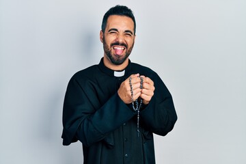 Sticker - Handsome hispanic priest man with beard praying holding catholic rosary sticking tongue out happy with funny expression.