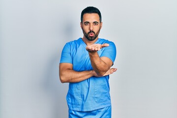 Wall Mural - Handsome hispanic man with beard wearing blue male nurse uniform looking at the camera blowing a kiss with hand on air being lovely and sexy. love expression.