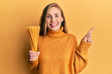 Poster - Young blonde woman holding dry spaghetti smiling happy pointing with hand and finger to the side