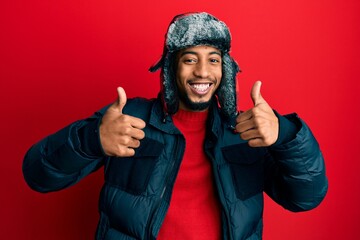 Young african american man with beard wearing winter hat and coat success sign doing positive gesture with hand, thumbs up smiling and happy. cheerful expression and winner gesture.