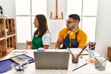 Poster - Young hispanic couple using laptop sitting on the table at art studio looking to side, relax profile pose with natural face with confident smile.