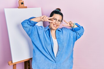 Poster - Young hispanic woman standing by painter easel stand doing peace symbol with fingers over face, smiling cheerful showing victory