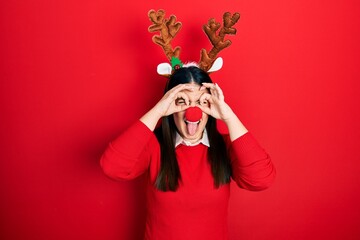 Sticker - Young hispanic woman wearing deer christmas hat and red nose doing ok gesture like binoculars sticking tongue out, eyes looking through fingers. crazy expression.