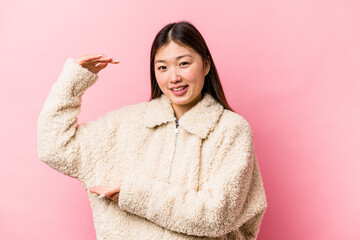 Wall Mural - Young Chinese woman isolated on pink background holding something little with forefingers, smiling and confident.