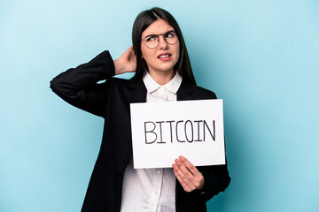 Young caucasian business woman holding a bitcoin placard isolated on blue background touching back of head, thinking and making a choice.