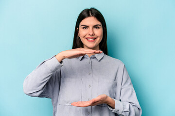 Wall Mural - Young caucasian woman isolated on blue background holding something with both hands, product presentation.