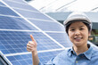 © Bond_JP - Close-up portrait with copy space of a confident Asian woman wearing white safety hat, smiling, posing thumb up, standing outdoor near solar panels. Female engineer and sustainable energy concept.