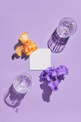two glasse with water and card note with iris flowers on pastel lilac background. summer refreshment