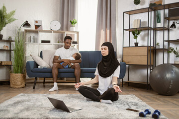 Arabian muslim woman in hijab meditating on mat in the living room using laptop as a tutorial for yoga fitness at home while her african husband puts little son to sleep while sitting on the couch