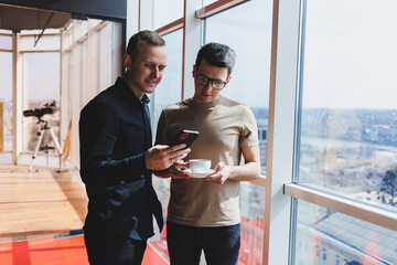 Two male businessman managers stand near the window in a modern office and discuss a new project. Two men with glasses are talking about a new business.