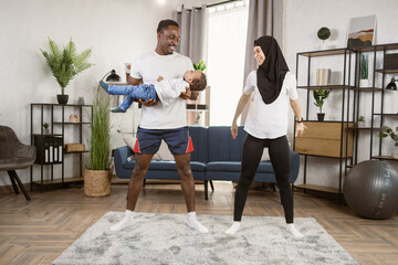 Sport, fitness, lifestyle and people concept. Smiling african man with a multiracial boy in his hands and muslim woman exercising and doing squats at home.