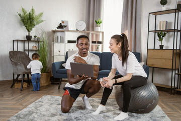 Wall Mural - Young caucasian woman in black sports leggings and a white t-shirt sitting on fit ball drinking water while african husband shows online lesson on laptop on the background of bright modern apartment.