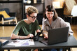 © AnnaStills - Young businesswoman explaining online work on laptop to her colleague while they sitting at table at office