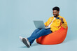Full body young man of African American ethnicity in yellow shirt sit in bag chair use laptop computer credit bank card shopping online order delivery isolated on plain pastel light blue background