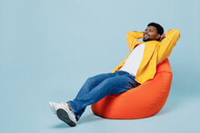 Full Body Smiling Happy Young Man Of African American Ethnicity Wear Yellow Shirt Sit In Bag Chair Look Camera Hold Hands Behind Neck Isolated On Plain Pastel Light Blue Background Studio Portrait