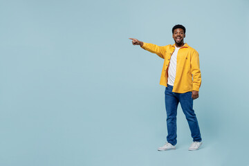 Wall Mural - Full body young amazed man of African American ethnicity 20s wear yellow shirt walking going point index finger aside on workspace area isolated on plain pastel light blue background studio portrait