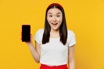 Wall Mural - Cheerful young girl woman of Asian ethnicity 20s years old wears white t-shirt hold in hand use mobile cell phone with blank screen workspace area isolated on plain yellow background studio portrait.