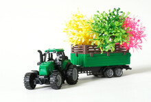 Green Toy Tractor Transports Green, Pink And Yellow Spring Seedlings In A Trailer. White Background. Copy Space. Selective Focus.