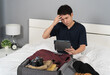 © geargodz - stressed man using tablet and packing clothes into suitcase on a bed, holiday travel concept