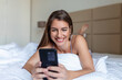 © Graphicroyalty - Smiling woman looking at mobile phone while laying on white bed. Happy brunette young woman using cellphone at home. Beautiful girl surfing the net on smartphone