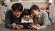 © PRPicturesProduction - Happy Asian girl lying on floor with her father and mother and having fun with online educational games on touchpad. Cheerful parents point at screen while giving advice to daughter