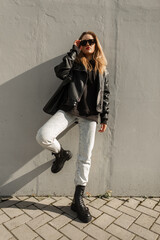 Wall Mural - Cool fashion young beautiful woman with trendy clothes with leather jacket, pullover, jeans and shoes with bag stands and poses near a gray wall on the street. Spring, fashion and style