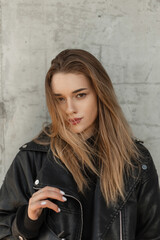 Wall Mural - Fashionable urban portrait of young beautiful woman with hairstyle in black fashion leather rock jacket with bag stands near a gray concrete wall on the street