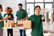© Syda Productions - charity, donation and volunteering concept - happy smiling female volunteer with food in box pointing to camera over international group of people at distribution or refugee assistance center
