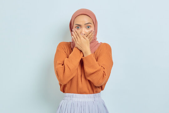 Shocked young Asian woman in brown sweater and hijab covering mouth with hand isolated on white background