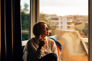 woman sitting on chair drinking a glass of wine and looking outside the window at sunset