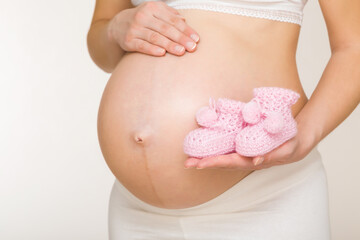 Wall Mural - Young adult pregnant woman with big naked belly on light gray background. Hand holding and showing pink soft knitted socks for future newborn. Closeup. Preparing clothes for baby in pregnancy time.