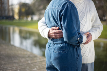 Elderly couple spending time together in park. Man in white sweater and woman in denim jumpsuit standing near pond in spring, hugging and holding hands. Relations, active rest of aged people concept